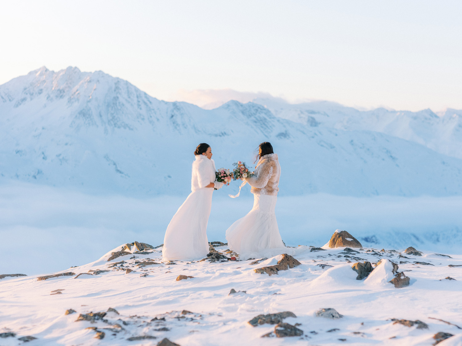 Alaska elopements, two brides on a snowy mountaintop looking at each other, bridal bouquet in hand, wearing faux fur coats