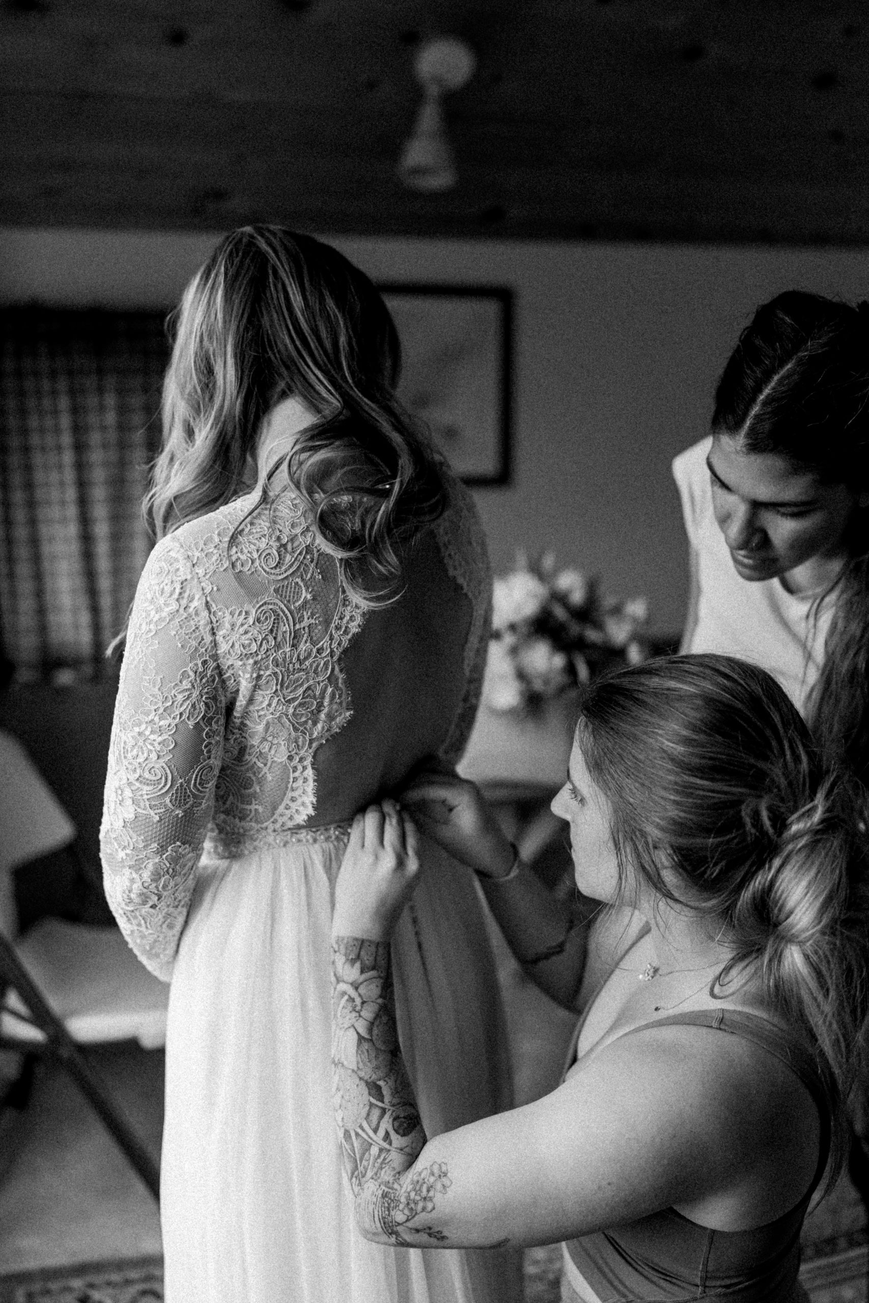 Black and white film photo of a bride's back, her friend buttoning up the back of her wedding gown.